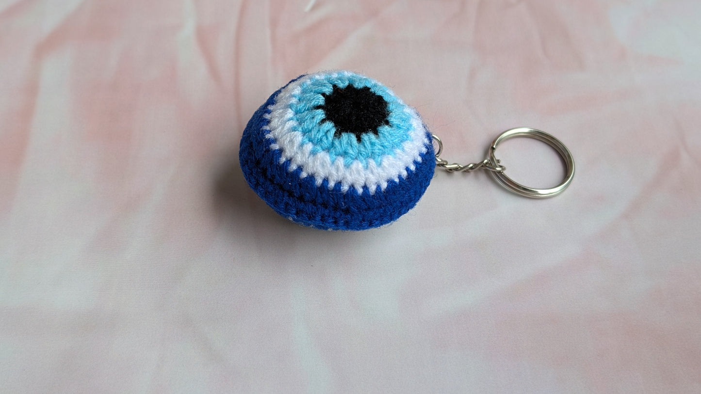 A crocheted evil eye keychain featuring blue, white, and black stripes with a silver metal key ring, resting on a light pink textured fabric background.
