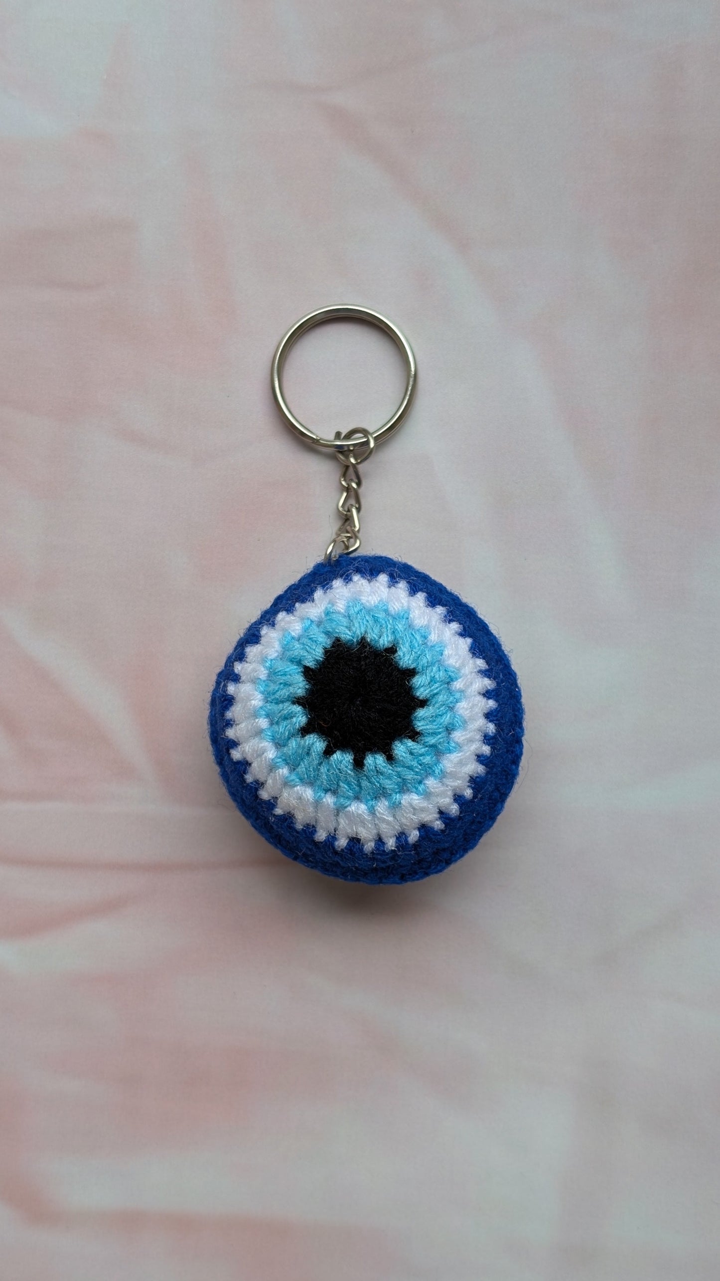 A crocheted evil eye keychain with concentric rings of blue, white, and light blue, featuring a black center and a silver metal key ring, shown on a soft pink fabric background.